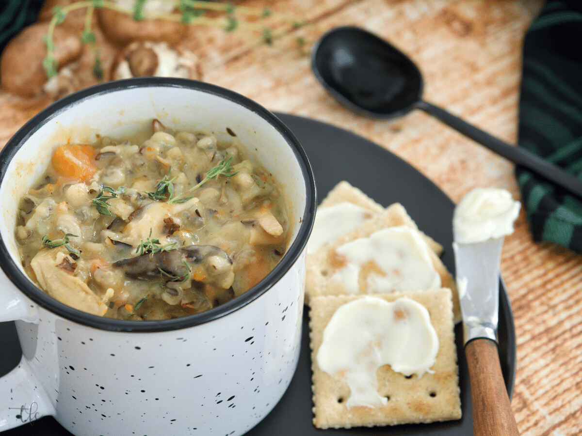 Speckled Mug filled with Instant Pot chicken and wild rice soup, spoon on the side, buttered saltines on a plate with mushrooms in background.