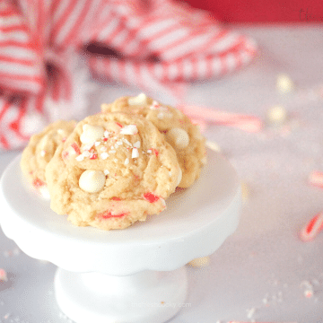 White Chocolate Peppermint Cookies on white pedestal with candy canes and white chocolate chips behind.