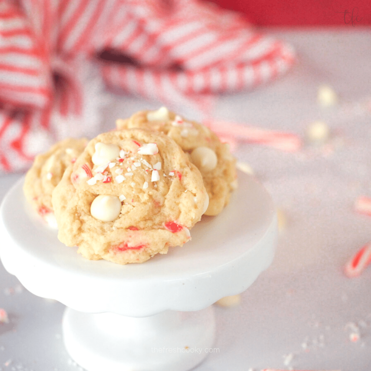 White Chocolate Peppermint Cookies on white pedestal with candy canes and white chocolate chips behind. 