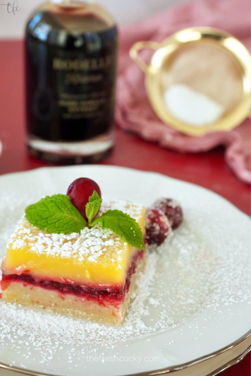 Cranberry lemon bar on plate dusted with powdered sugar, Rodelle Reserve vanilla in background.