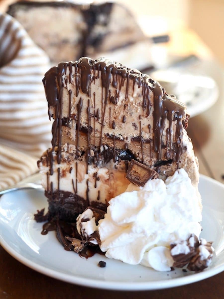 Slice of tall mile high mud pie ice cream cake with whipped cream and chocolate drizzle.