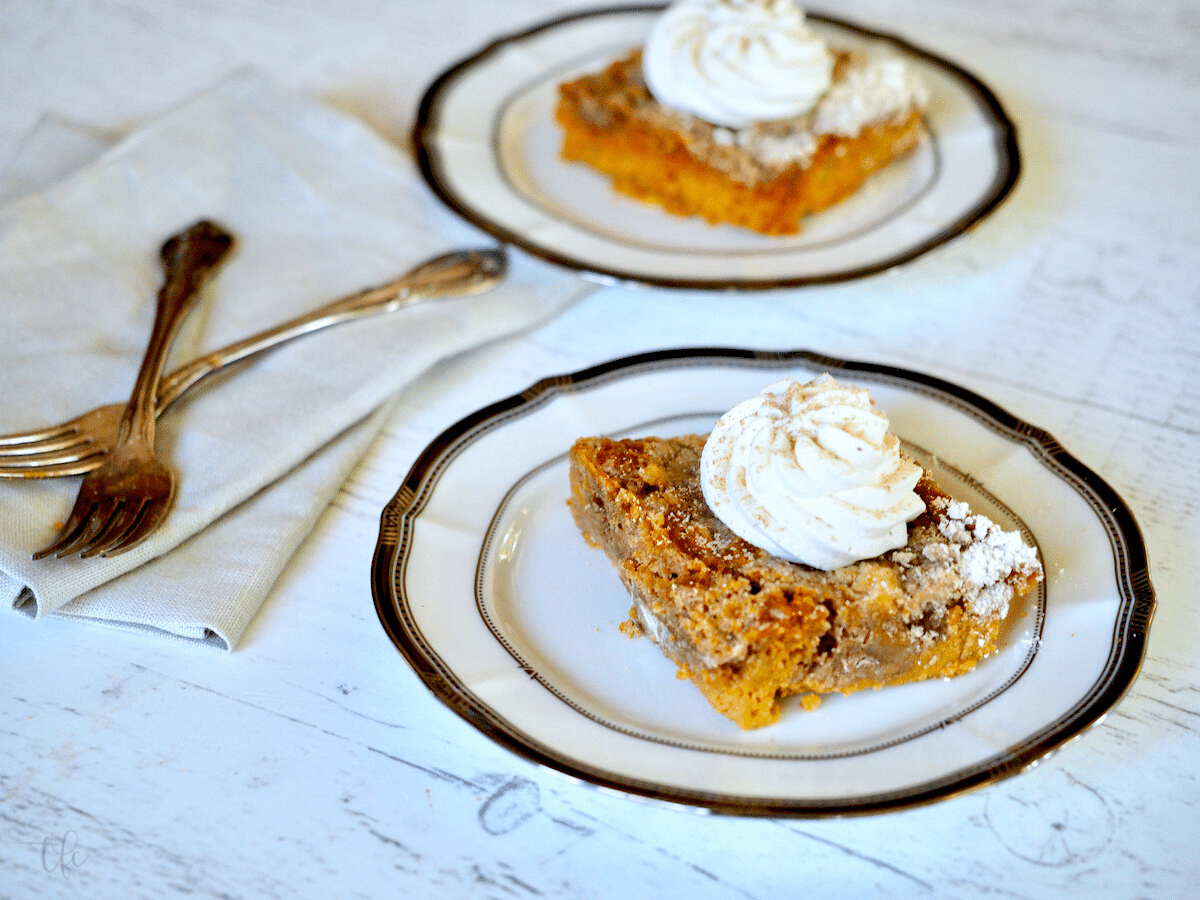 Two slices of pumpkin pie dump cake on pretty plates with forks and napkins, pie is topped with whipped cream.
