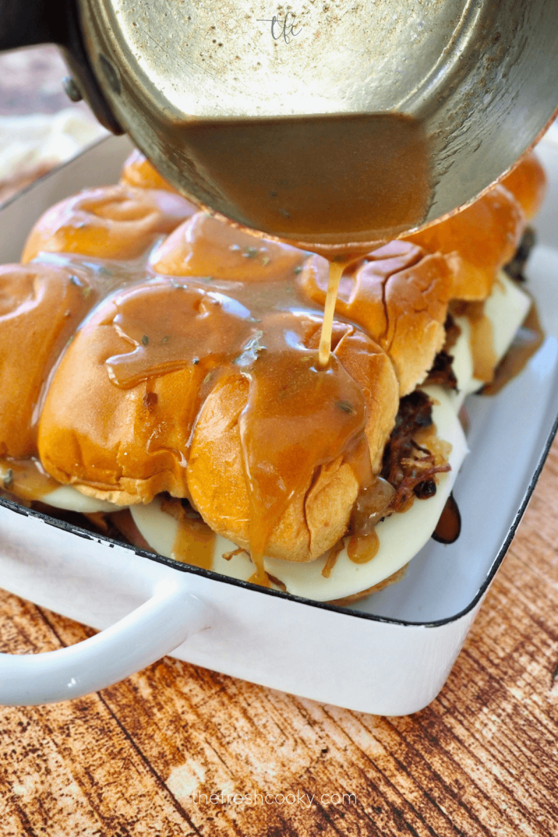 Pouring savory butter sauce over the tops of the roast beef sliders. 