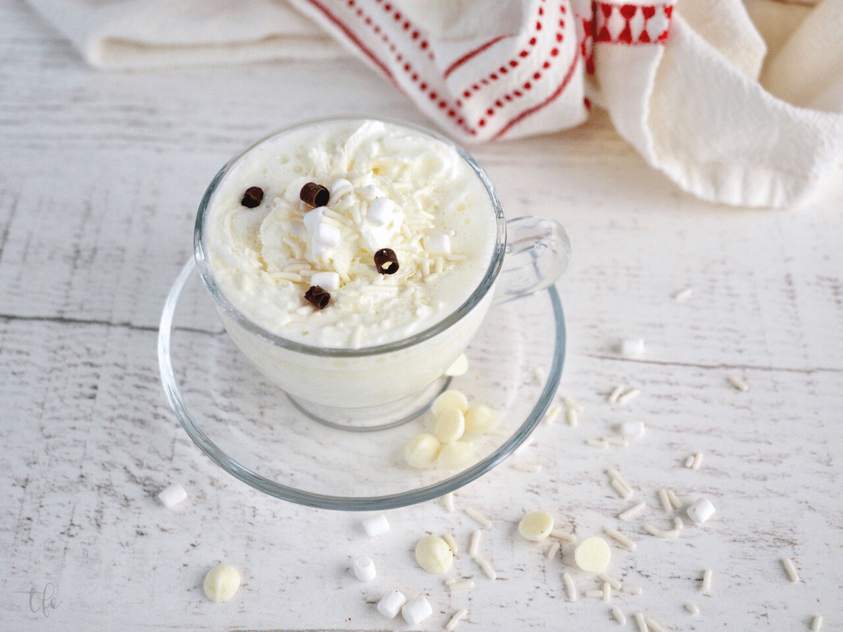 Starbucks white hot chocolate in small glass mug with plate, sprinkled with white and dark chocolate curls, whipped cream and sprinkles.