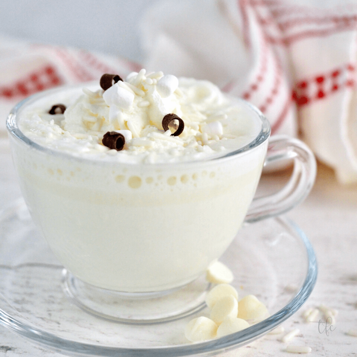 Starbucks Copycat White Hot Chocolate square image in clear glass teacup mug with whipped cream, white chocolate chips, chocolate curls and mimi marshmallows.