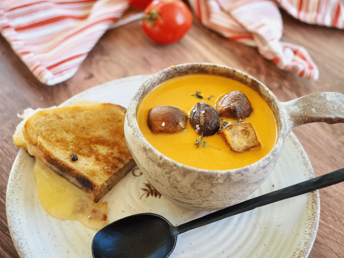Creamy tomato bisque recipe in stoneware bowl with black spoon and a melty grilled cheese on a plate.