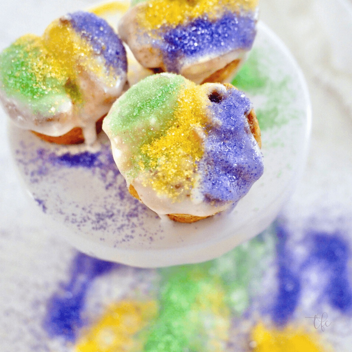 Easy Mini King Cake Bites on a small pedestal with Mardis Gras colored sugars sprinkled around.