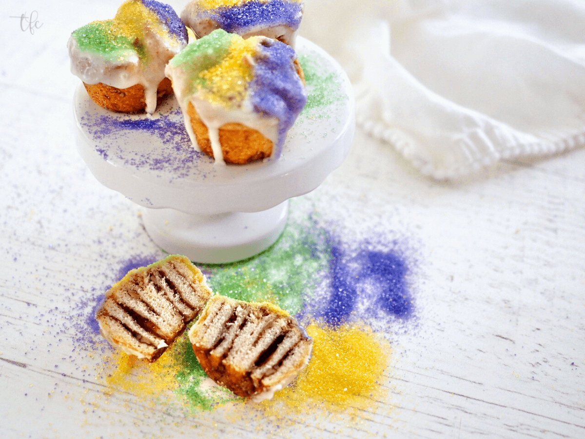 Easy Mini King Cakes on pedestal with one king cake bite cut in half revealing cinnamon sugar insides.