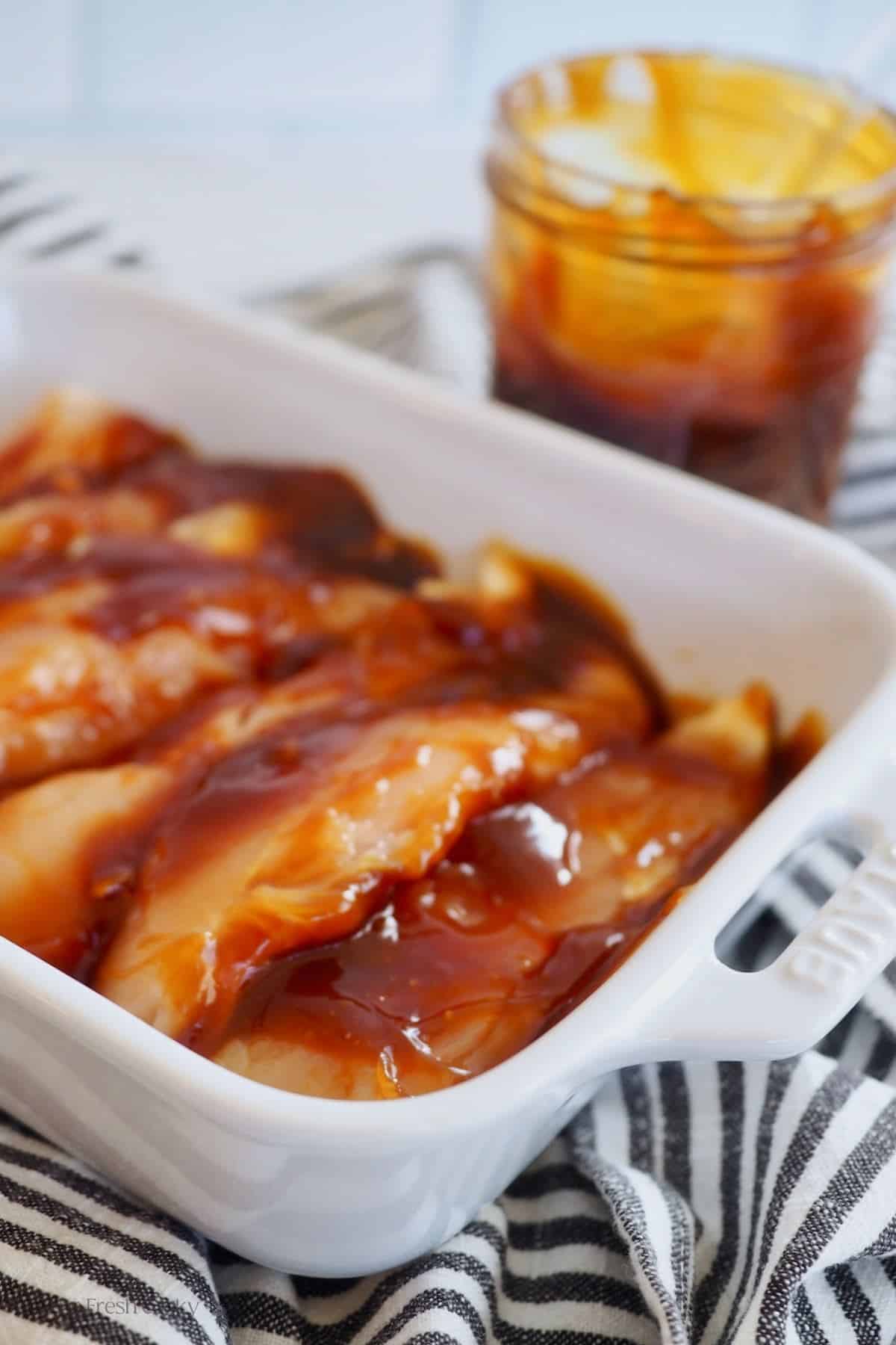 Staub white casserole dish with raw chicken marinating in teriyaki sauce with jar behind.