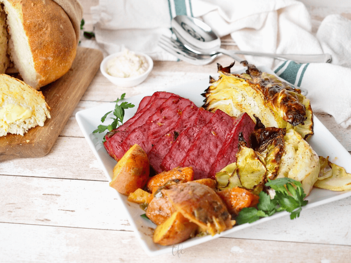 Horizontal image of baked corned beef recipe on platter with loaf of Irish Soda Bread behind.