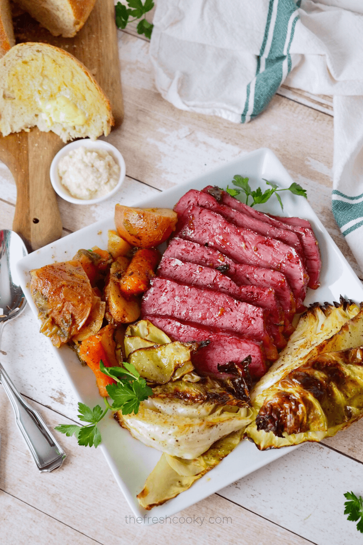 Baked Corned Beef recipe on plate with potatoes, carrots and roasted cabbage with bread in background.