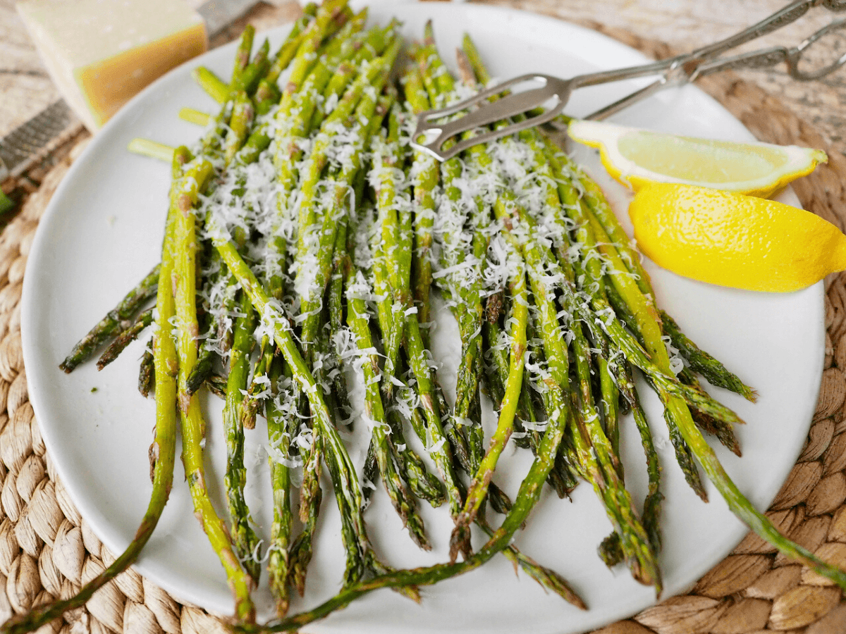 Roasted Air Fryer Asparagus on a plate with lemon wedges and sprinkles of parmesan with tongs to serve.