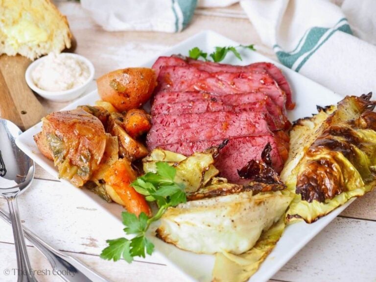 Baked Corned Beef recipe on plate with potatoes, carrots and roasted cabbage with bread in background.