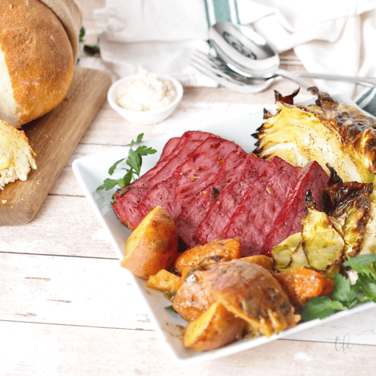 Square baked corned beef recipe with meat on platter surrounded by roasted cabbage, potatoes and carrots and a loaf of bread in background.