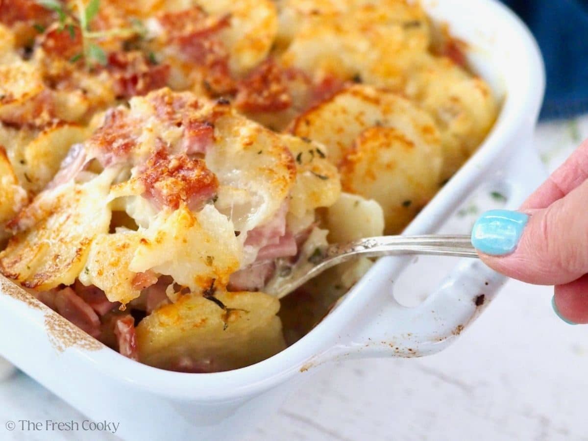 Hand serving with a pretty silver spoon, cheesy scalloped potatoes with ham.