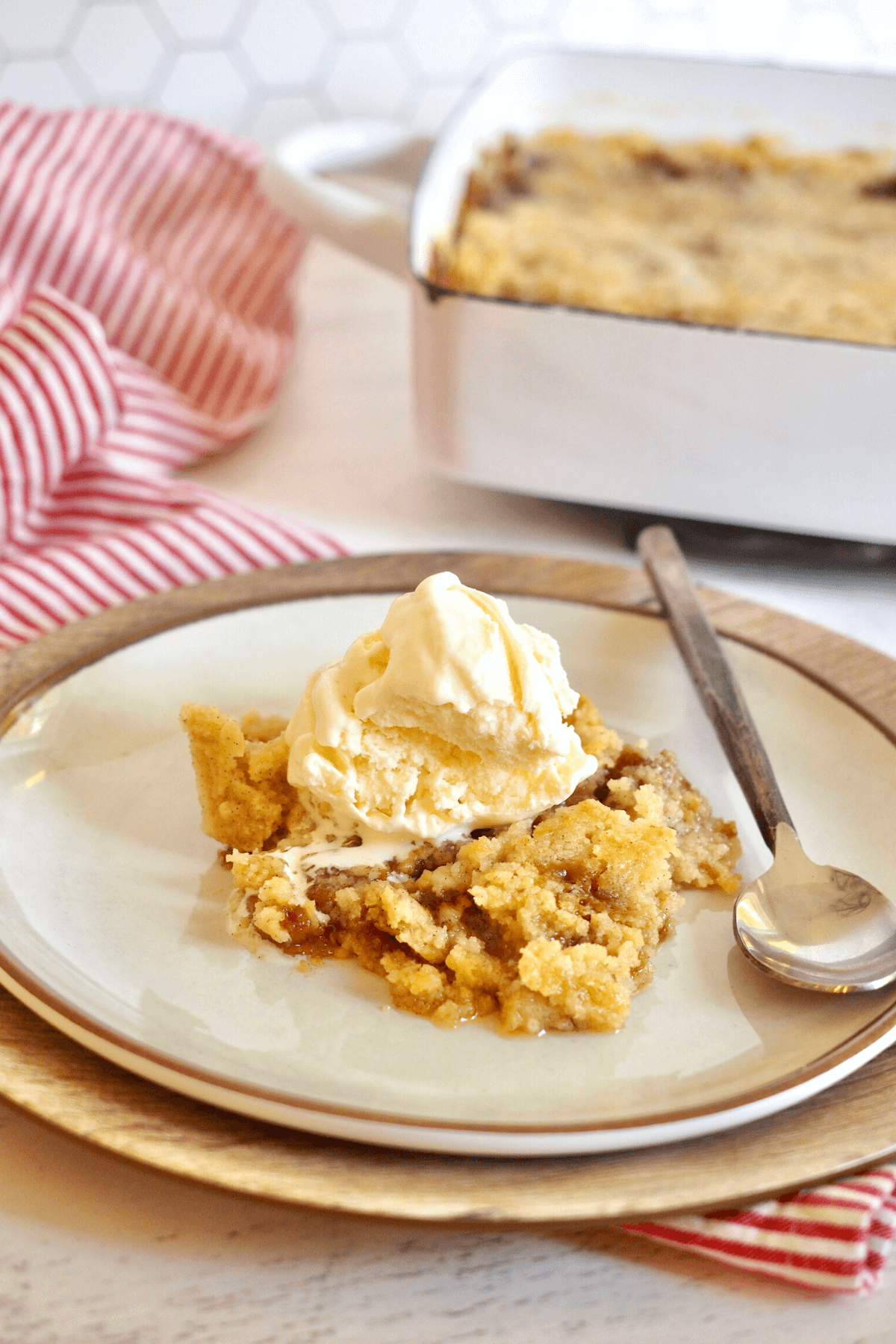Caramel Apple Dump Cake slice on a plate with a spoon ready to dig in, scoop of vanilla ice cream on top and pan of dump cake mixture behind.
