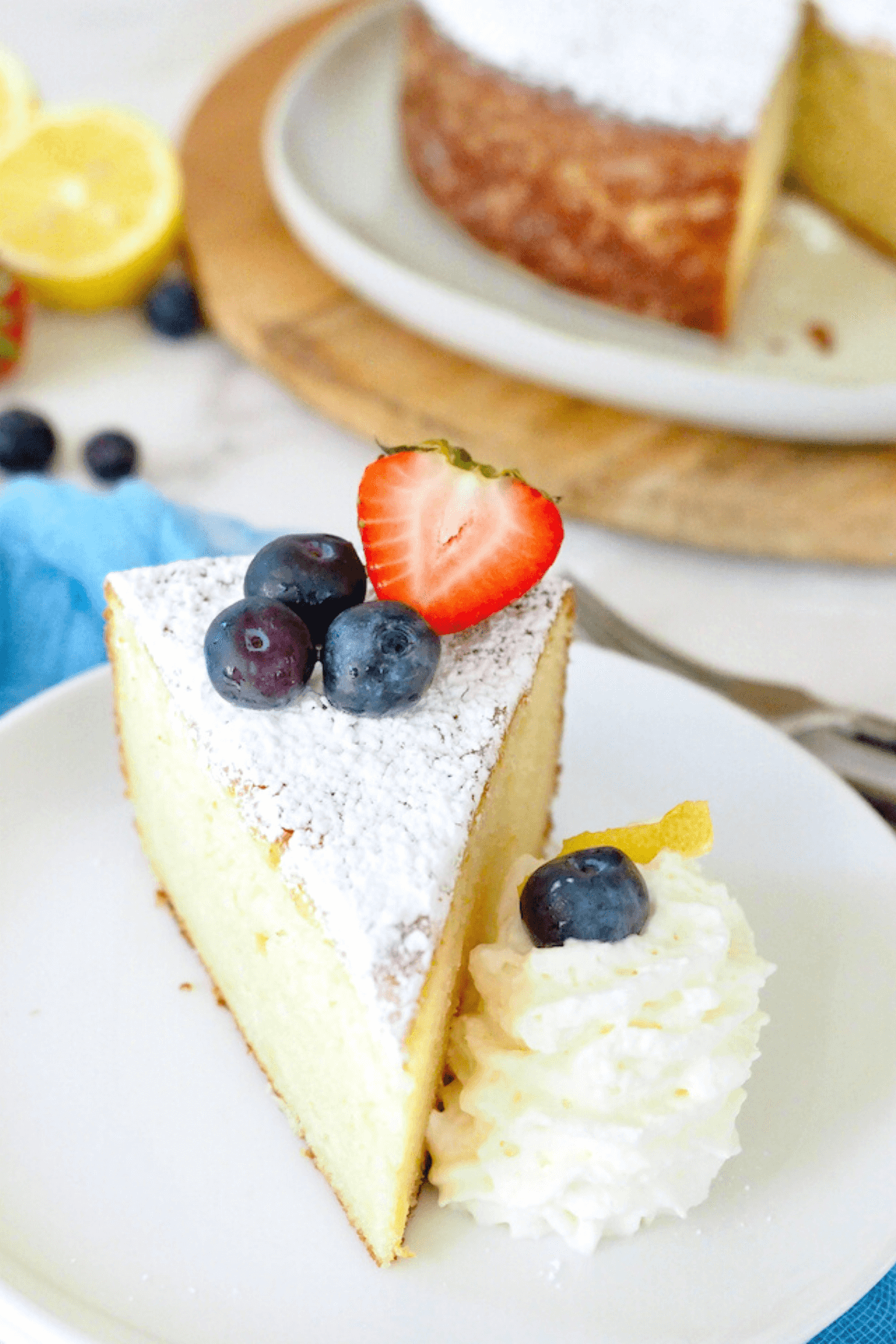 Slice of lemon ricotta cake on plate with whole cake in background, slice is topped with blueberries and strawberries along with a dollop of whipped cream.