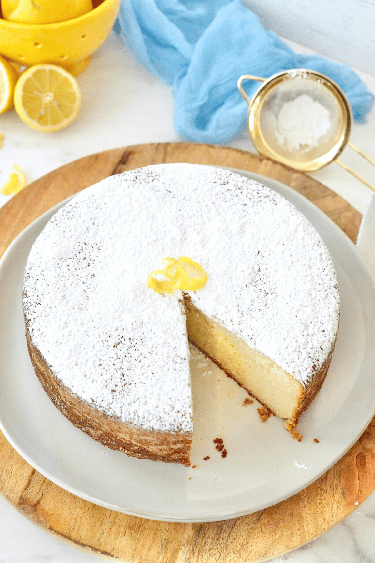 Lemon Ricotta Cake with slice removed and fresh lemons and powdered sugar in sifter nearby. 