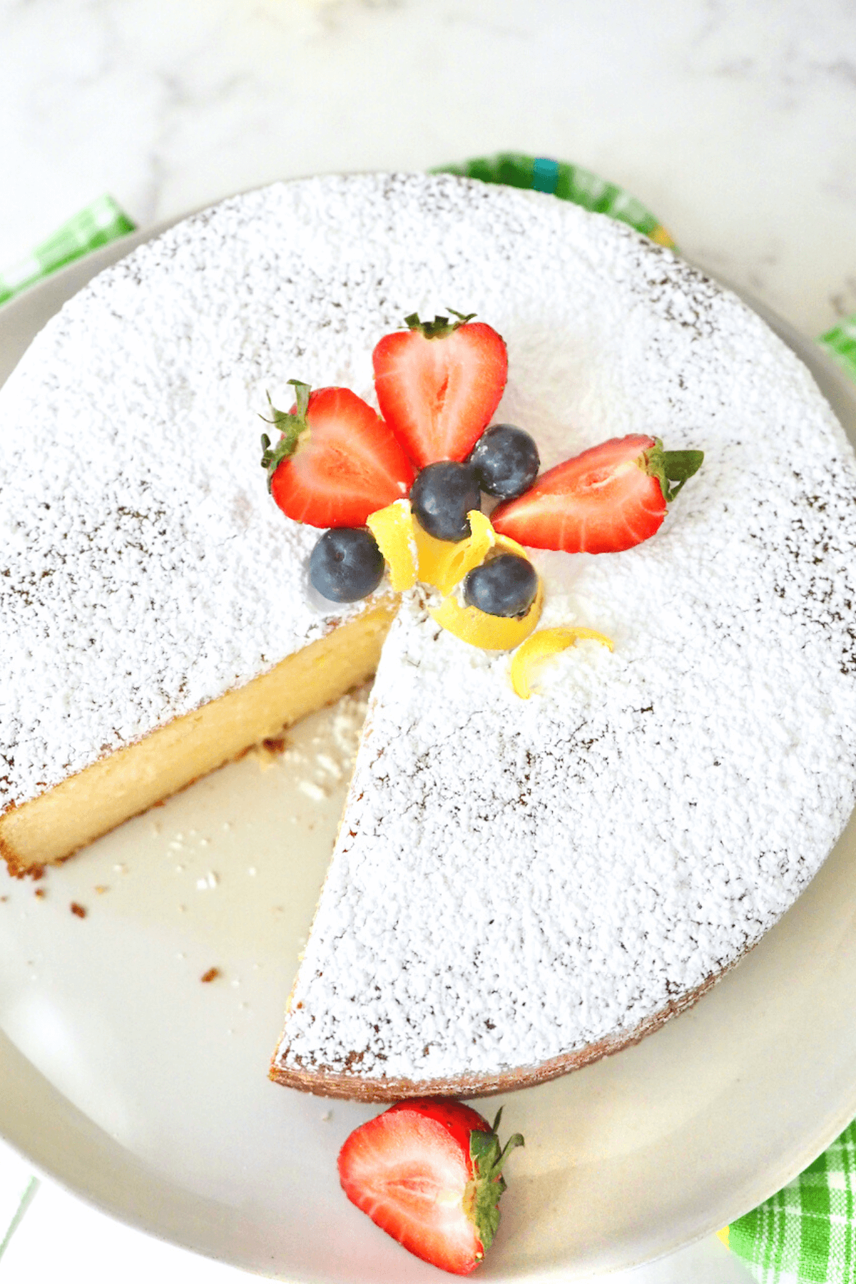 Italian Lemon Ricotta Cake Recipe with slice removed and decorated with fresh strawberries, lemon rind and blueberries.