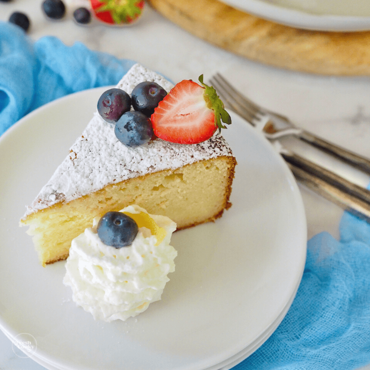 Lemon ricotta cake recipe slice on white plate with forks behind, cake is decorated with whipped cream and berries.