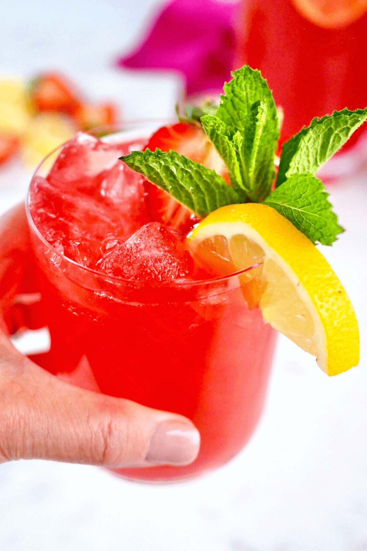 Hand holding glass filled with bright pink lemonade vodka drinks, garnished with lemon, strawberry and mint.
