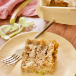 Slice of Rhubarb Cake recipe on a bamboo plate with a rustic fork and ribbons of rhubarb in background.