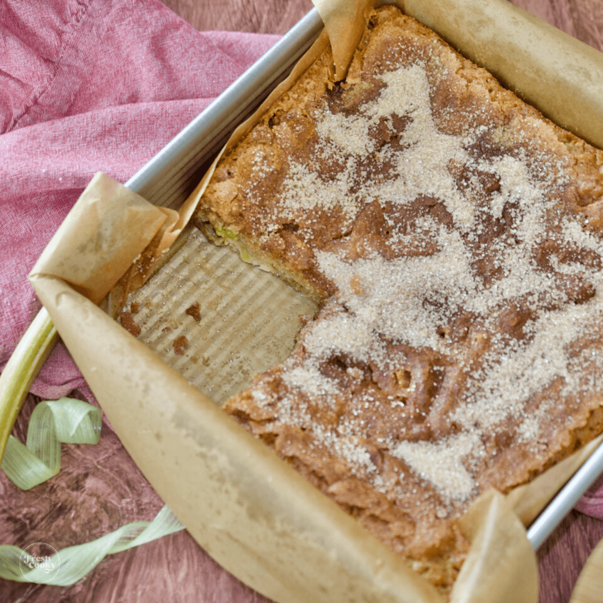 Square pan filled with rhubarb coffee cake with one slice missing, ribbons of rhubarb and a pink napkin.