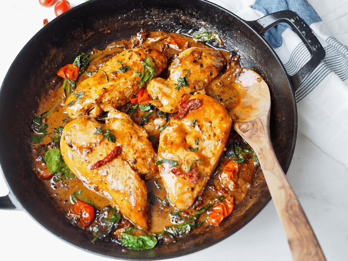 Creamy Tuscan Chicken in skillet with wooden spoon surrounded by cream sauce and sun-dried tomatoes.