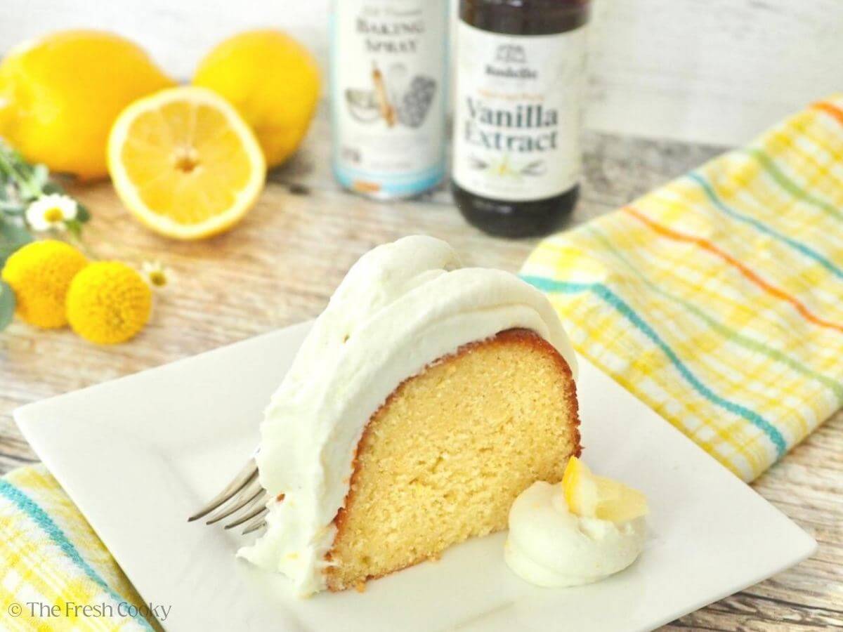 A slice of lemon bundt cake with lemons, flowers, baking spray, and Rodelle vanilla extract behind.