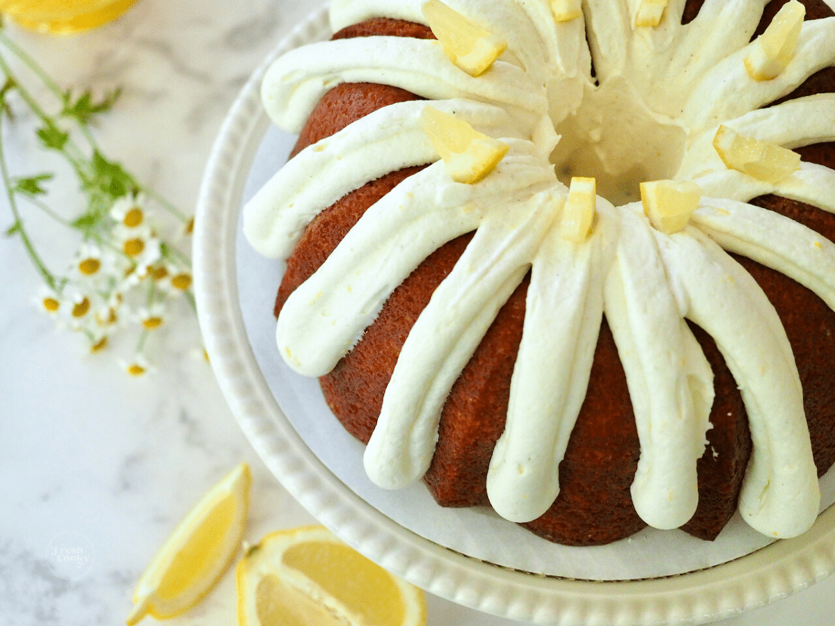 Lemon bundt cake top down shot with whole bundt on cake platter with lemons and flowers.