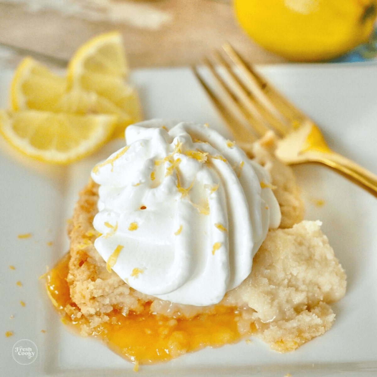 Slice of lemon dump cake on plate topped with whipped cream, lemon wedges and gold fork in background.
