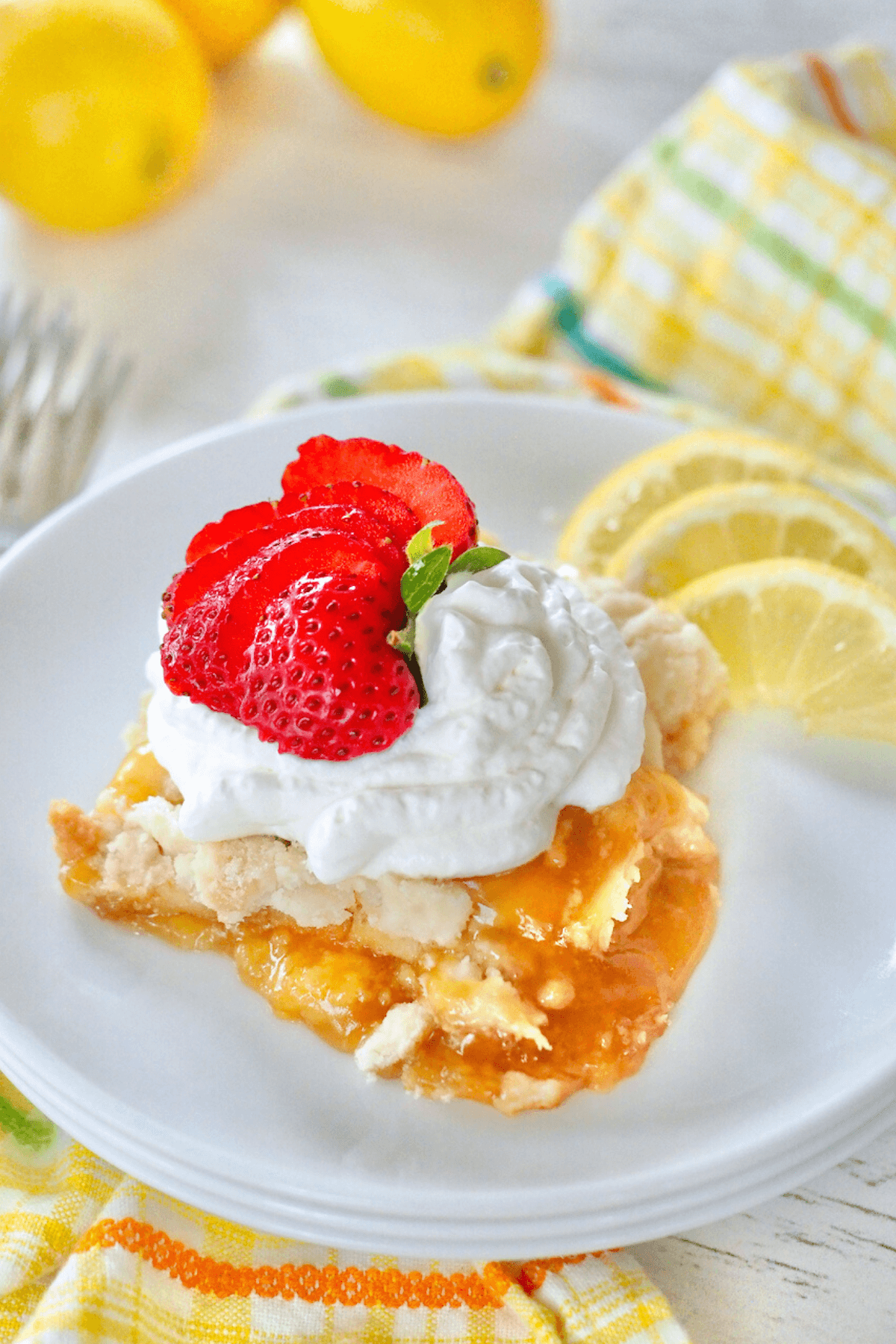 Lemon dump cake recipe sliced and on plate topped with whipped cream and strawberries, with lemon slices in background.