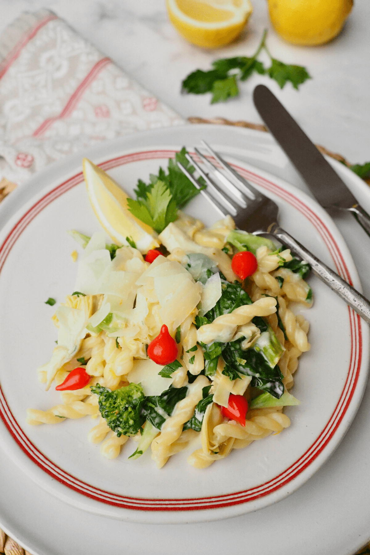 Lemon garlic pasta recipe on plate with pops of sweet peppers, spinach, artichoke hearts, lemon wedge and garnished with shaved parmesan.