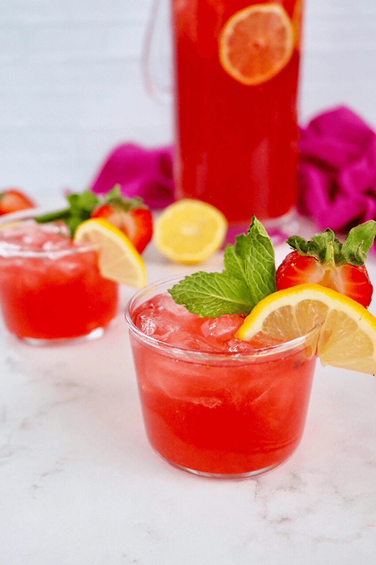 Pink lemonade in short bodega glasses with pitcher of pink vodka lemonade behind.