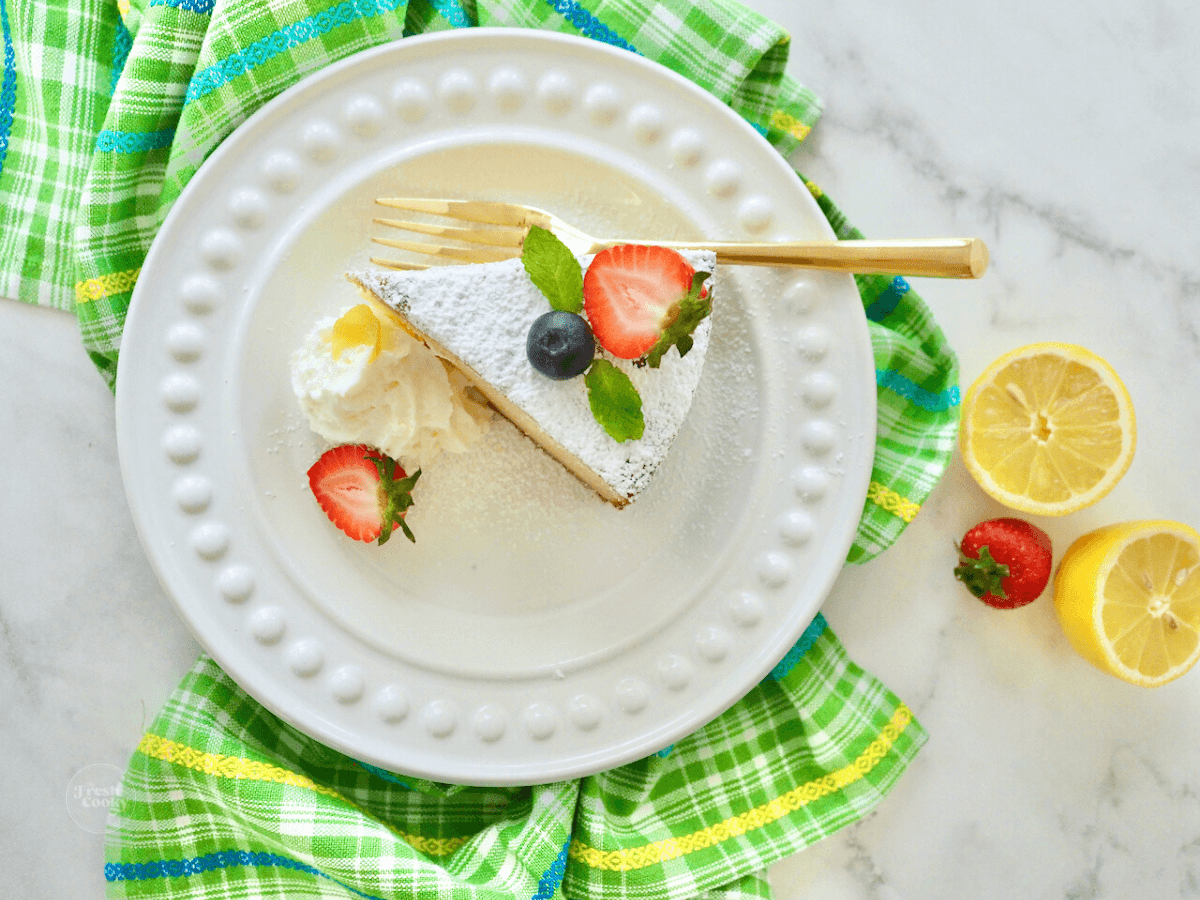 Top down shot of wedge of lemon ricotta cake slice on plate with gold fork and whipped cream. 