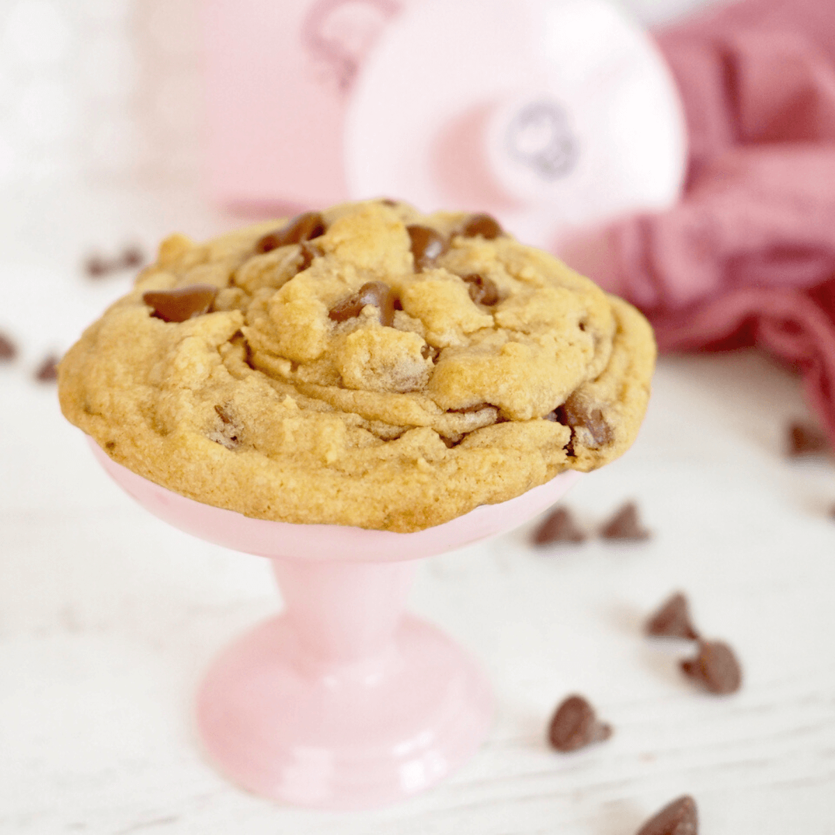 Crumbl copycat chcoolate chip cookie recipe with giant cookie on pink pedestal and chocolate chips laying about.
