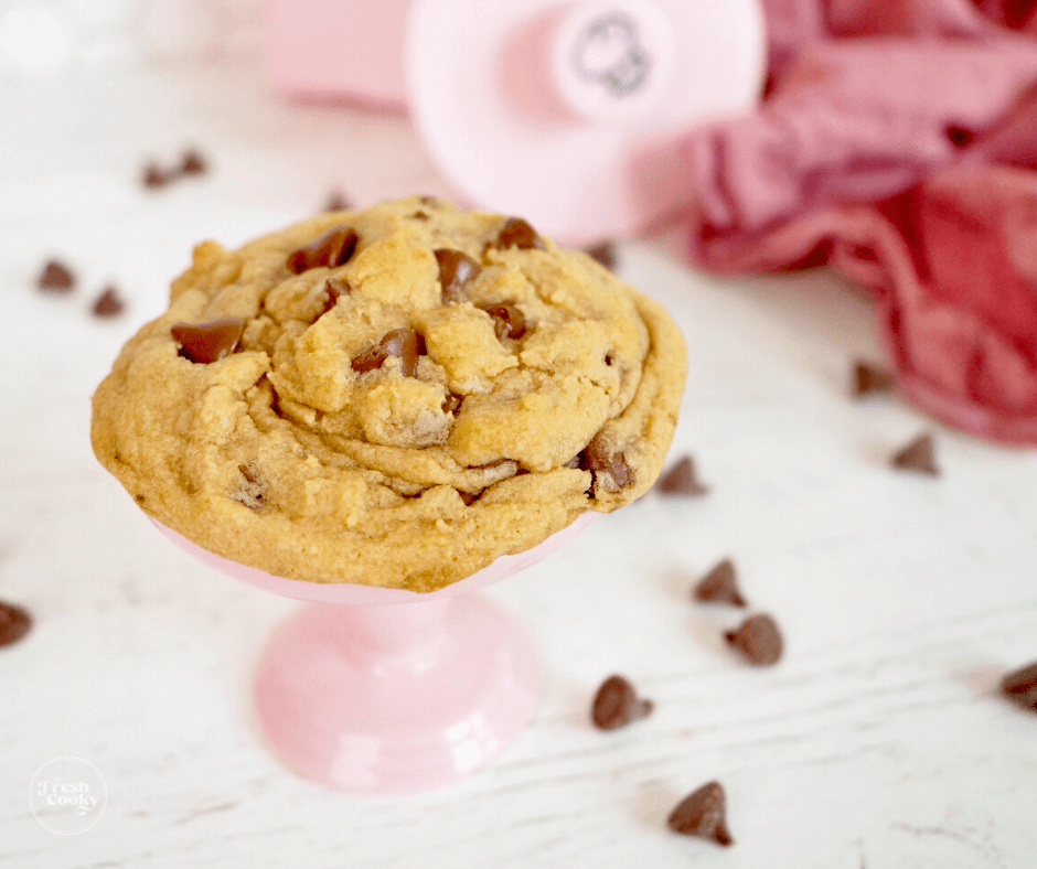 Copycat Crumbl chocolate chip cookie on pink pedestal with crumbl cookie cutter in background and chocolate chips laying about.