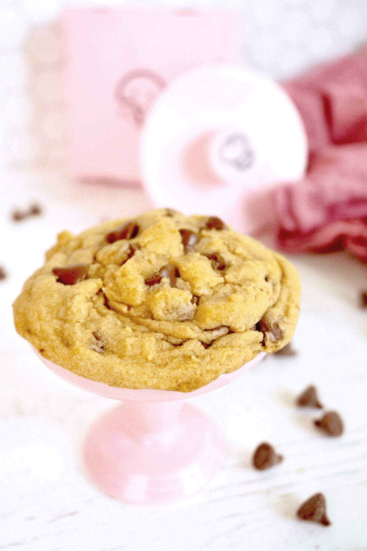 Crumbl Cookie chocolate chip on pink pedestal with crumbl cookie box and cookie cutter behind, scattered milk chocolate chips around cookie.