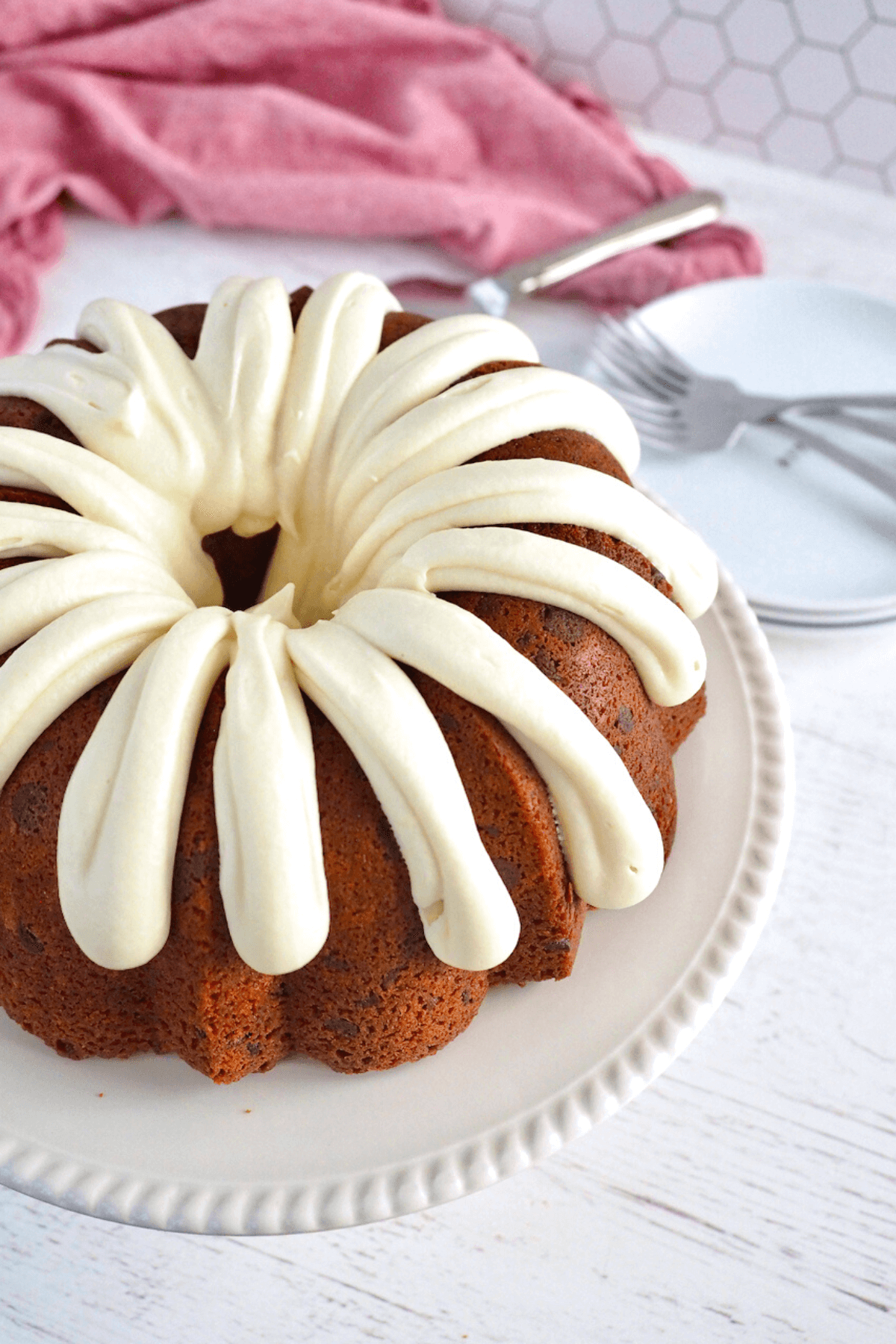 Frosted red velvet bundt cake frosted with thick rich fingers of cream cheese frosting, on a pretty cream cake plate with forks behind.