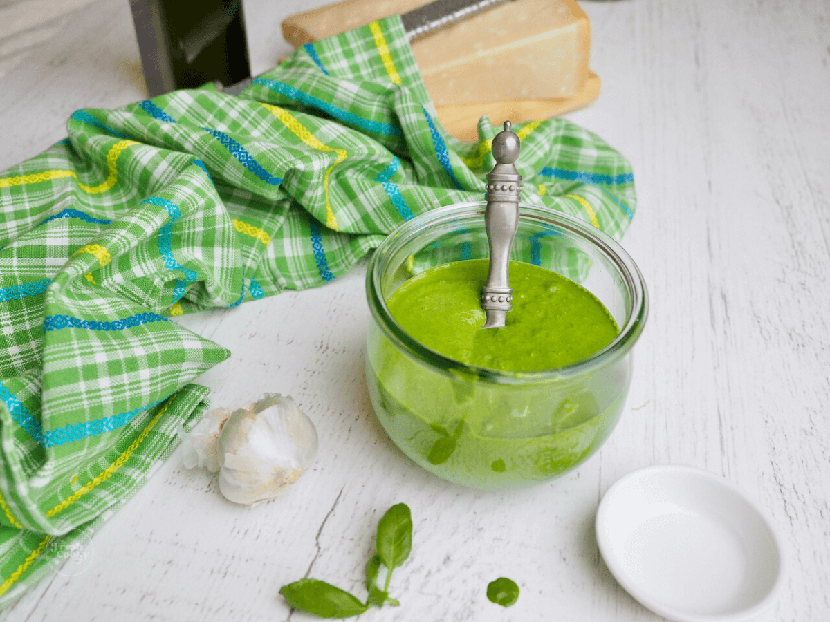 How to make pesto without nuts image of pesto on table in a jar with a spoon and fresh garlic, basil and a hunk of parmesan cheese in the background.