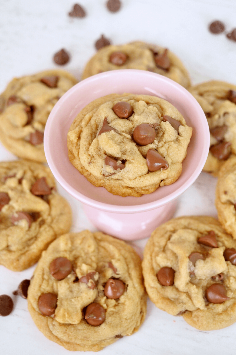 Mini Crumbl chocolate chip cookies with one cookie in a pink pedestal and other mini cookies surrounding. 