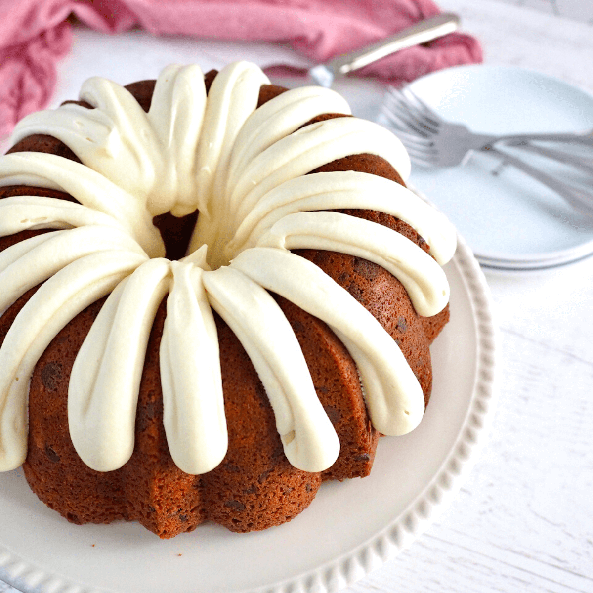 Red Velvet Bundt Cake on cream plate with thick fingers of Cream Cheese Frosting, pink napkin behind with two white plates and forks. 