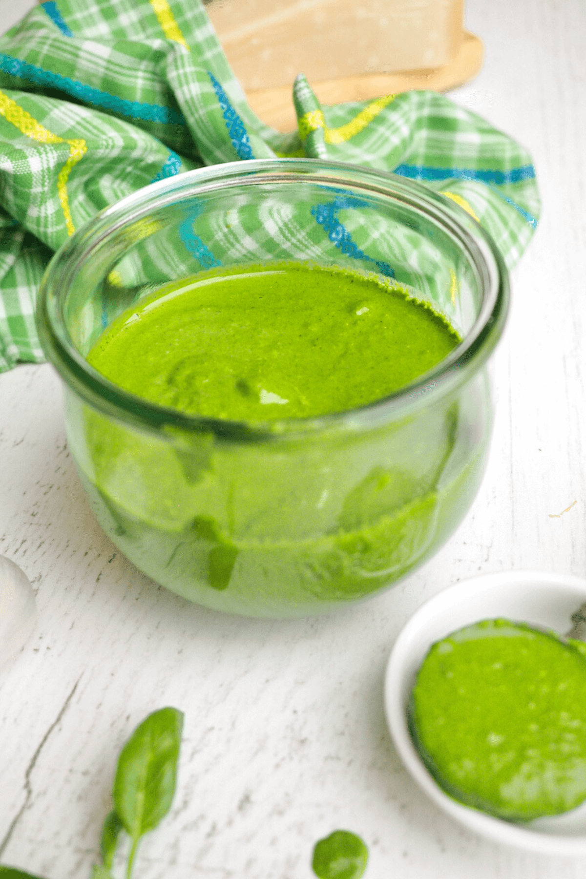 Jar filled with bright green spinach and basil pesto without nuts with spoonful in small dish nearby and fresh basil leaves on table.