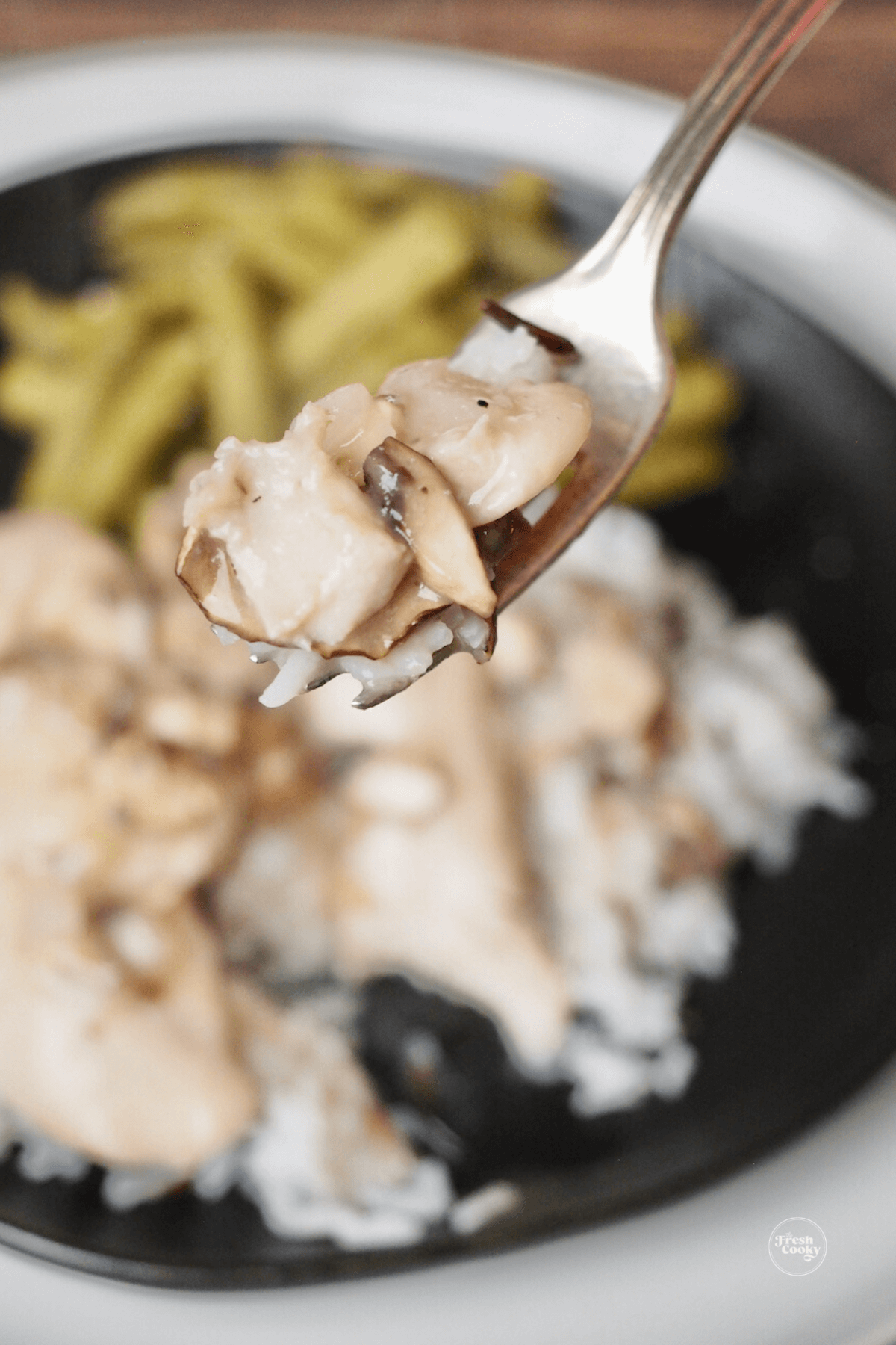 Fork with tender piece of chicken, mushrooms and gravy with plate of chicken and rice in background.