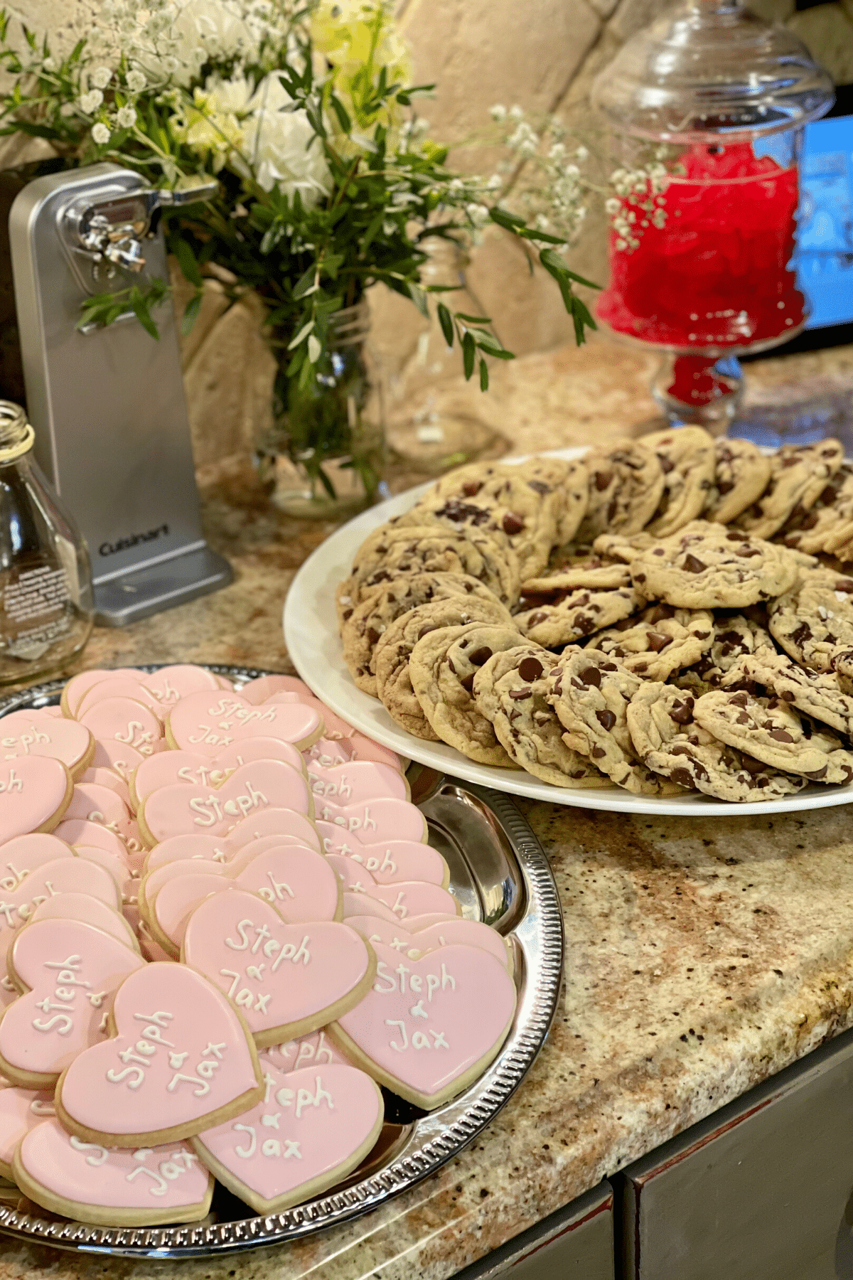 Countertop with trays of Crumbl Chocolate chip cookies along with decorated sugar cookies for the bride and groom. 