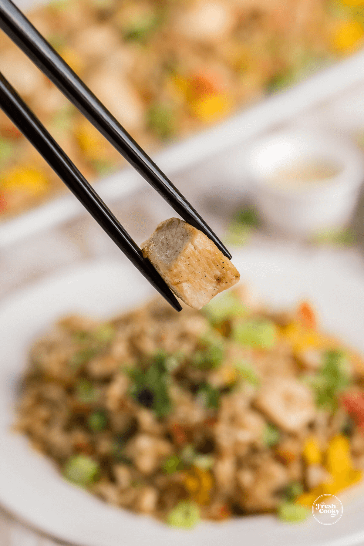 Chopsticks holding piece of chicken with fried rice in background.