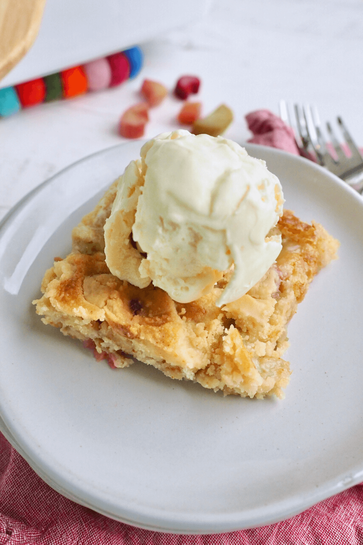 Slice of rhubarb dump cake on a plate with a scoop of vanilla ice cream, forks in background along with pieces of rhubarb.
