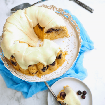 Blueberry Pancake Bundt Cake Recipe with cake frosted and on pedestal with slice removed and on plate.
