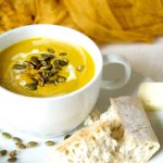 Panera Autumn squash soup in white soup bowl topped with roasted pepitas and served with French bread.