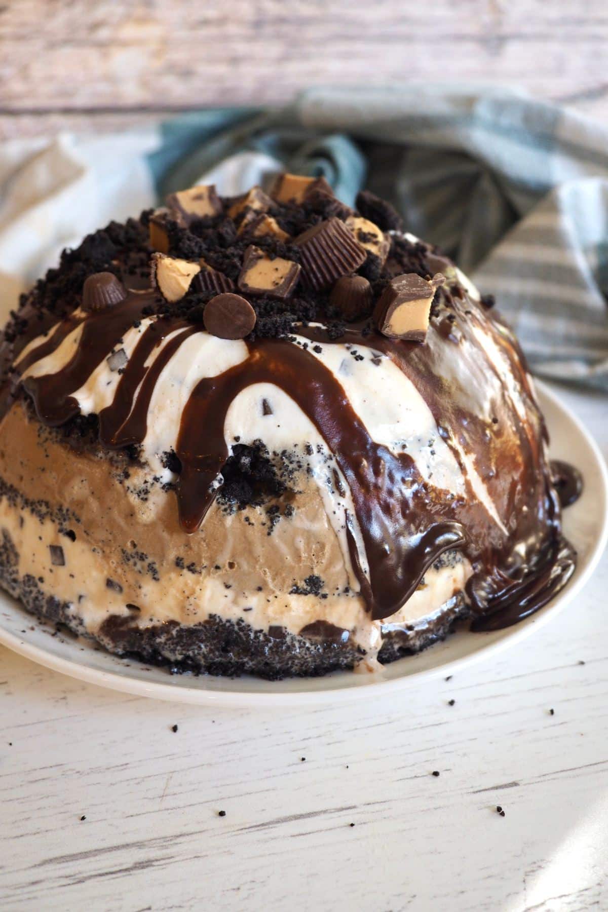 Beautiful mud pie layered ice cream cake on plate, drizzled with fudge and topped with peanut butter cups.